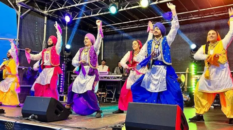 Men and woman in traditional Sikh dress - of loose trousers, a top and waistcoats - dance on stage. Each outfit is a different colour - of blue, green, gold, orange and pink - with men wearing turbans. The dancers have one arm raised in the air with lights shining behind them.