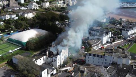 An aerial view of smoke billowing from the derelict hotel. The building is white and the roof is black with smoke.