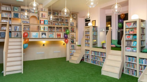 A children's library with green carpet, cushions, steps, ballons and books on shelves.