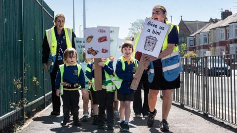 Young boys and girls in their blue and yellow school uniform hold up placards saying "Give us a bin!" as they march on a pavement, between railings on both sides. They are accompanied by two female school staff with hi-vis jackets.