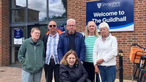 6 people stand outside a brick building with blue and white signage saying 'Shropshire Council' 'Welcome to the Guildhall' . A person in a green jacket and glasses stands on the outside, next to a man in sunglasses and a striped blue and white shirt. Next to him is a man with white hair and glasses in a blue coat, holding on to the wheelchair of the lady at the bottom of the picture who has brown hair and a navy jacket on. Next to them are two women, one is blonde with a green and white striped top and a lady with glasses and a white top. They all look unhappy.