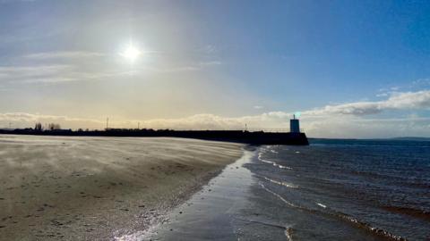 Sunshine and mainly blue sky over the coast in Nairn, Highland. Sandy shore with pier in the background.