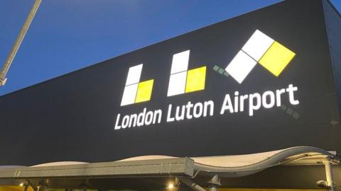 The front of London Luton Airport. A large black building with a large illuminated LLA and "London Luton Airport" written on it in white font.