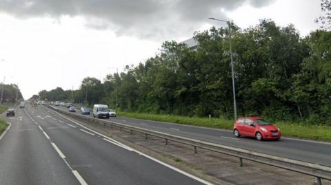 Cars drive along a motorway. There are two lanes on each side of the central reservation, trees can be seen on the far right. 