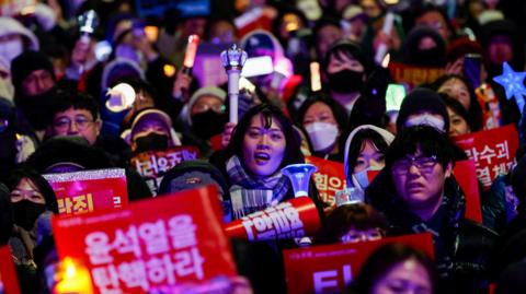 A close up of a sea of faces at a protest in South Korea. Several people are carrying signs or waving candles and flashlights.