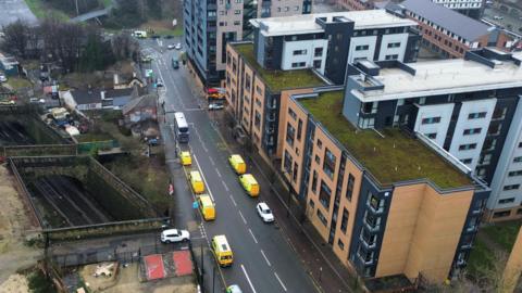 An aerial view of an urban area featuring a wide road running through the centre of the scene. Along this road, several yellow emergency vehicles, likely ambulances, are lined up in a row, parked on one side of the street. A red emergency vehicle, possibly a fire engine, is also visible further up the road near an intersection. A white bus is stopped in the middle of the road, and a few other cars are nearby.