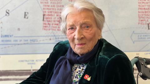 Christian wearing a velvet jacket and a scarf with white hair and blue eyes smiling at the camera. She is in a wheelchair and you can see a map in the background but it is not in focus.