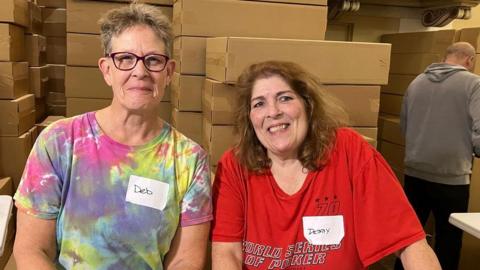 Two women are standing together in a room full of cardboard boxes. The woman on the left has short brown/grey hair and is wearing a tie-dye T-shirt with a name badge that reads Deb. The woman on the right has shoulder-length brown wavy hair and is wearing a red T-shirt with a name badge that reads Debby. Both are wearing colourful T-shirts and have name badges.