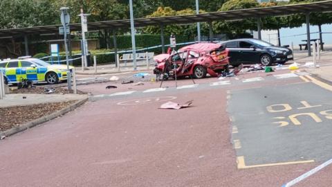 Mangled wreckage of red car surrounded with debris (middle) and police car (left) with a black car behind the crashed one