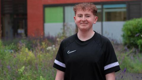 Harrison Sherwell, with short brown curly hair is wearing a black casual fit t-shirt with a thick white horizontal stripe on the sleeves. He is smiling, standing against a backdrop of wildflowers.