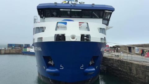 The image shows the front of a large blue-and-white freight ship moored at a harbour, with ropes tied to the quay and a few people standing near the dockside in the background.