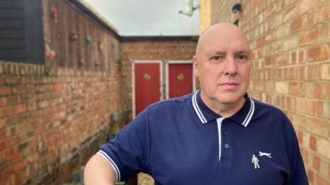 A man with a shaved head, dressed in a blue polo shirt, stands in a narrow yard between two red-brick walls. Two red wooden doors with white frames can be seen in the background.