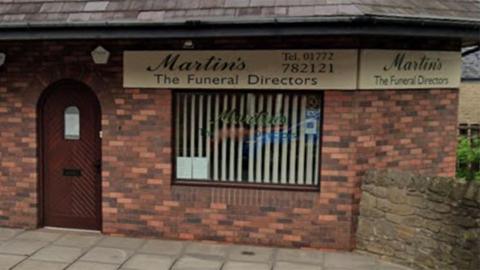 Streetview image of the funeral directors in Longridge. A red brick building with a brown arched door to the left and a window to the right with a vertical cream blind.