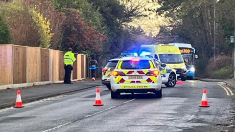 Two police cars and an ambulance blocking a road. There are three small orange and white traffic cones blocking the road in the foreground. A police officer in yellow high-vis coat is stood on the path on the left of the image. A bus can be seen facing the camera in the background behind the emergency vehicles.
