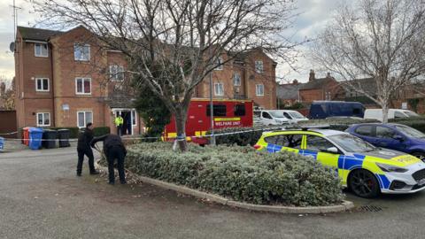 Two police officers wearing black uniforms search a bush in a car park outside a block of flats. Police tape blocks the front of the block and car park. A police car is parked and there is also a red welfare unit in the car park.