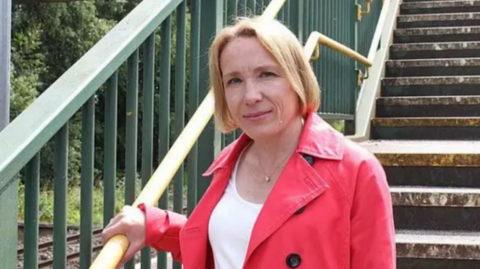 A woman with blonde hair, a white top and red jacked standing on some steps and holding green metal railings