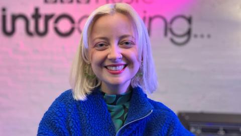 Caswell, inside the BBC Music Introducing studio, wearing a bright blue fleece zip jacket and vibrant red lipstick