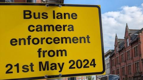 A yellow sign reads "Bus lane camera enforcement from 21st May 2024" on Corporation St in Preston.