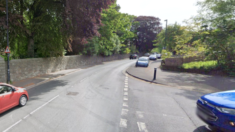 Blackberry Hill in Stapleton, Bristol. Cars are driving past trees and a stone wall