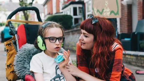 Mother Laura with her son Oscar. Oscar is in a wheelchair wearing ear defenders whilst his mum looks at him dotingly.