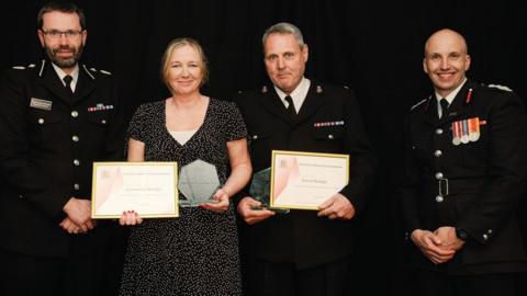 Three men in uniform with a woman in a dark top. Samantha and Dave Bridge are holding certificates