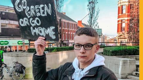 Izaak Cowell holding a sign "Go Green or Go 鶹Լ" while protesting in Preston