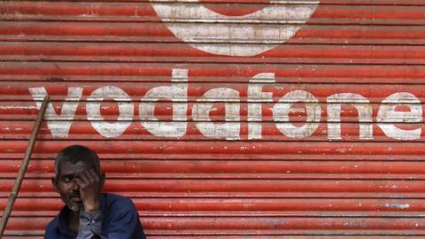 A man sits outside the downed shutters of a shop painted with a logo of Vodafone on its shutter in Mumbai, India on 24 February 2019