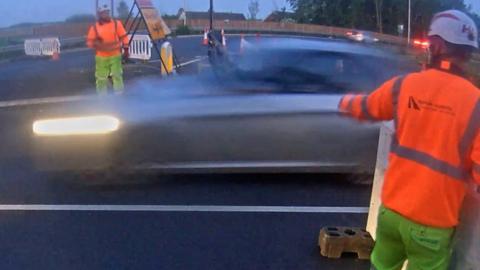 People in orange jackets standing beside a moving car