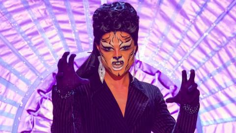 Gothy Kendoll on the Ru Paul's Drag Race UK runway in 2019. Gothy wears a black pinstripe suit and black wig and has painted her face to resemble a tiger. She holds her hands up gesticulating a roar.