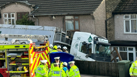 White lorry with smashed windscreen appears to be in contact with a row of houses after apparently leaving the road. Firefighters and police in hi-vis stand in the foreground on the road, with two fire engines visible