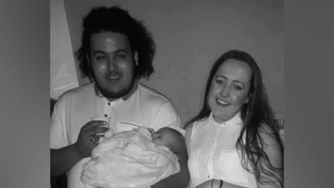 A black and white photo shows Brendan Ledgister holding his baby daughter and standing next to his partner Chelsey Berriman