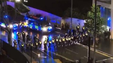 A line of more than 30 police officers moves towards crowds in Stratford, with two police vans following behind with their blue lights on.