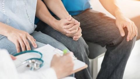 A couple hold hands as a doctor with a clipboard takes notes