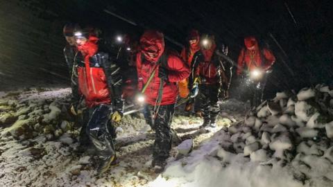 A group of volunteers in red and black waterproofs walk at night, lit by head torches, in sleet and snow