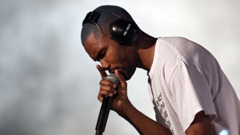 A side-shot photo of Frank Ocean looking down, holding a microphone close to his face, wearing a white anti-discrimination T-shirt while singing at a gig.