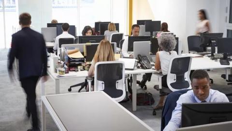 Staff sat at desks looking at computer monitors in an open-plan office as a man in a suit walks away from the camera