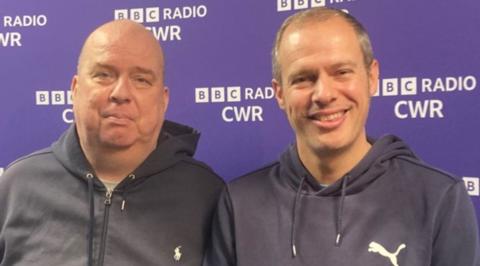 Neil Jeffs on the left is standing next to younger brother Paul, who is smiling at the camera. They are in front of 91ȱ Radio CWR branding