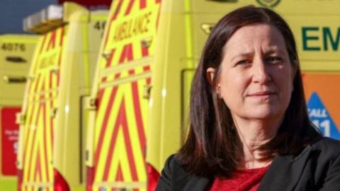 A woman with dark hair and a red top with a dark jacket with her arms folded in front of a row of three yellow and red striped ambulances