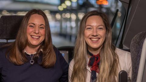 Two female drivers Jess and Emma are sitting in the cab of a train and smiling. They are both young with long hair and behind them you can see Newcastle station .