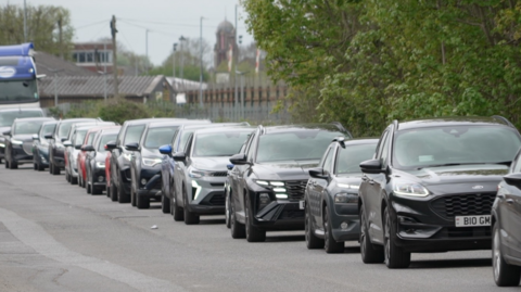 A queue of more than a dozen cars, followed by a large truck with blue livery, on a grey asphalt road lined by green trees. In the background, a wooden fence and the tops of buildings can be seen, along with the red and white raised barriers of a railway crossing.