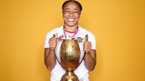 Sadia Kabeya holds the Women's World Cup trophy in front of a gold background, dressed in her England rugby kit