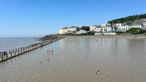 A large marine lake with people swimming and paddle boarding in it. The water is a murky brown colour, and the sky is blue, with old hotels and houses lining the promenade in the distance.