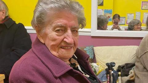 Margaret smiles into the camera. She's wearing a purple coat. A mirror behind her shows a young girl taking the photo. Margaret has short, white hair. 
