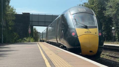 A train is pulling out of a small rural station and passing under a road bridge. The train is dark green with a yellow section on the front which has GWR printed on it.