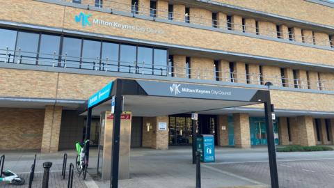 A multi-storey brick office building bearing the sign “MK Milton Keynes City Council,” viewed from street level on a clear day. The façade features repeating vertical window recesses and metal railings along each level. At ground level, a covered entrance canopy displays the same council branding, with signage, a bicycle, and a parked e‑scooter visible near the entrance.