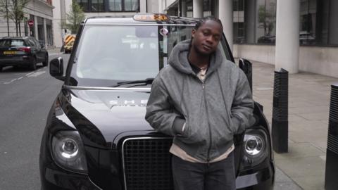 A man wearing a grey hoodie standing in front of a black cab