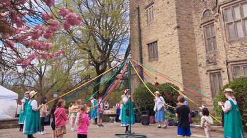 People in traditional green and blue costumes dancing around a maypole outside a large historic stone building, weaving colourful ribbons beneath blooming pink cherry trees on a bright spring day.