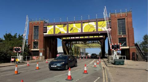 A road bridge with two red-brick towers and raised barriers under a clear blue sky. Several orange and white traffic cones guide vehicles through narrowed lanes as cars pass beneath the structure. The bridge features a large yellow decorative panel with overlapping circle patterns.