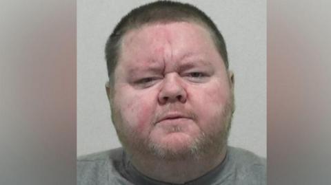 Mugshot of Mark Hogarth. He has a large round head with a stubbly fair beard and short dark hair. he is wearing a grey sweatshirt.