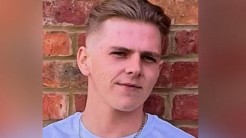A young man wearing a white t-shirt and silver chain stands in front of a brick wall. He is looking at the camera.